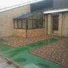 Green solid top in a pathway going towards a building surrounded by bricks and stones. The solid top has puddles from rain on them.